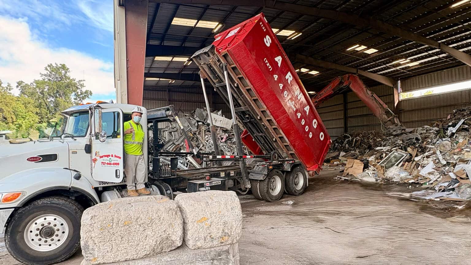 Red Sarnago roll-off container being tipped inside the covered transfer station, a sorted construction-debris pile behind and a worker in hi-vis beside the truck