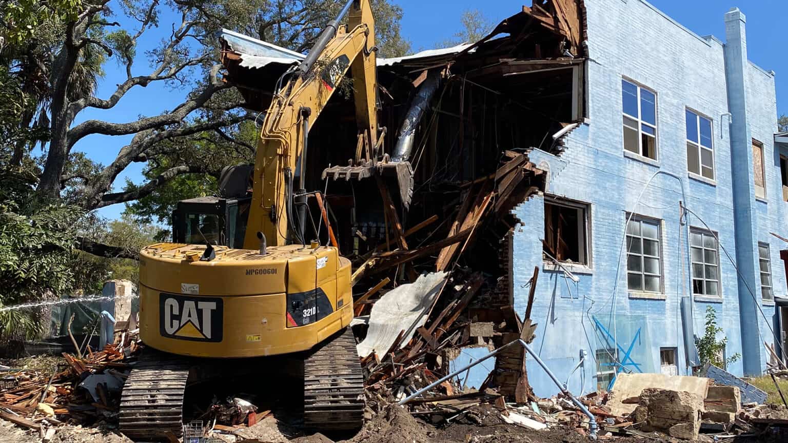 Excavator tearing into the upper story of a blue masonry building, roof and timbers coming down