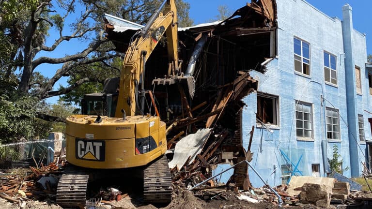 Excavator tearing into the upper story of a blue masonry house, roof and timbers coming down